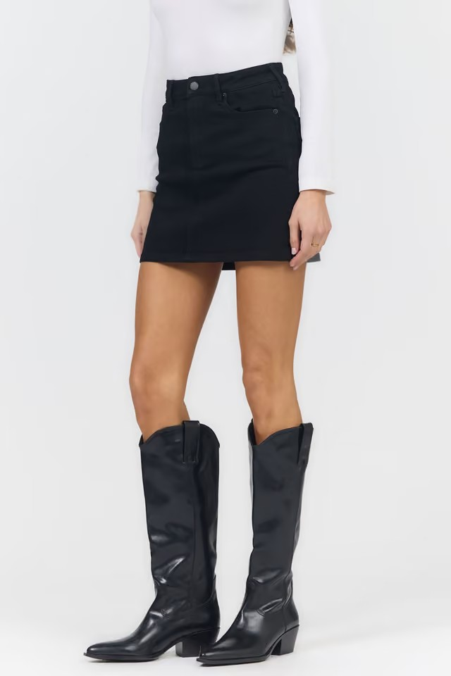 A model wearing a black denim mini skirt with a white long-sleeve top and black knee-high leather boots, shown from the waist down on a light gray studio background.