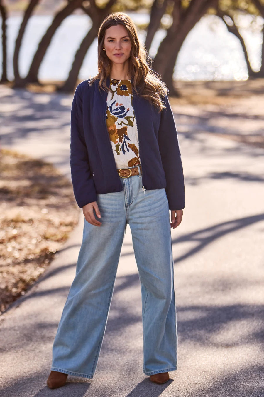 A woman stands on a sunlit paved path outdoors wearing a navy cardigan over a cream floral top, light blue wide-leg jeans, and brown shoes. Trees and blurred outdoor scenery are visible in the background.