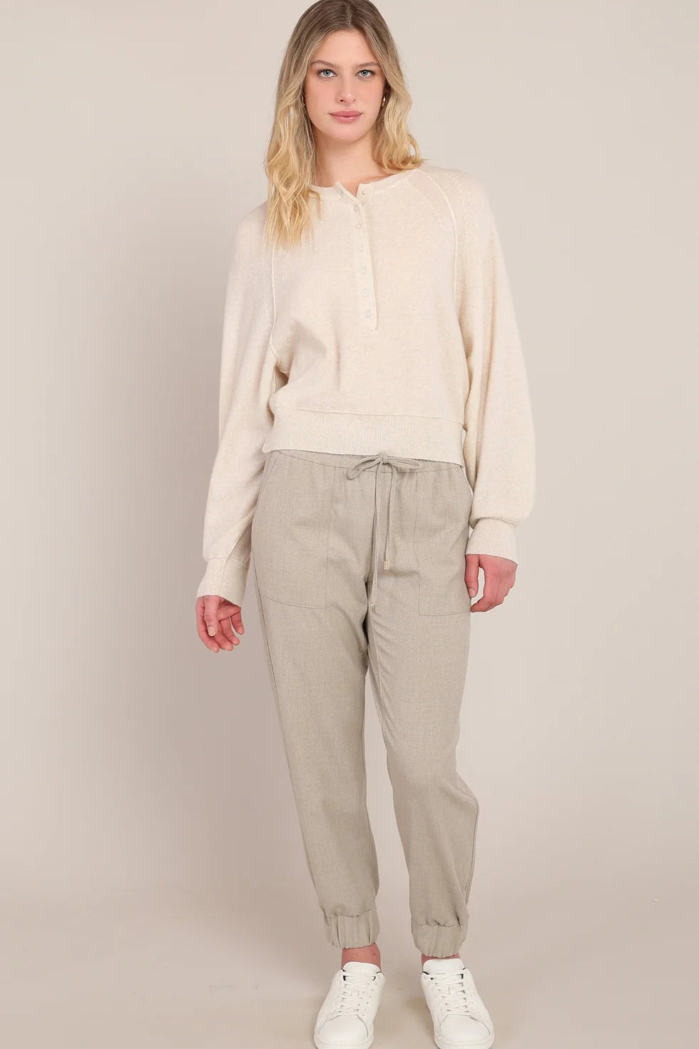 A woman modeling a cream-colored long-sleeve knit top and light beige drawstring trousers, paired with white sneakers, against a solid light beige studio background.