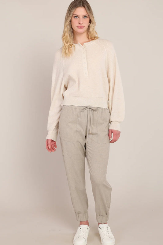 A woman modeling a cream-colored long-sleeve knit top and light beige drawstring trousers, paired with white sneakers, against a solid light beige studio background.