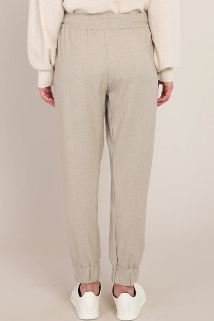 A person shown from the back wearing light beige jogger-style pants with an elastic waistband and cuffed ankles, paired with a cream sweater and white sneakers against a plain beige studio background.