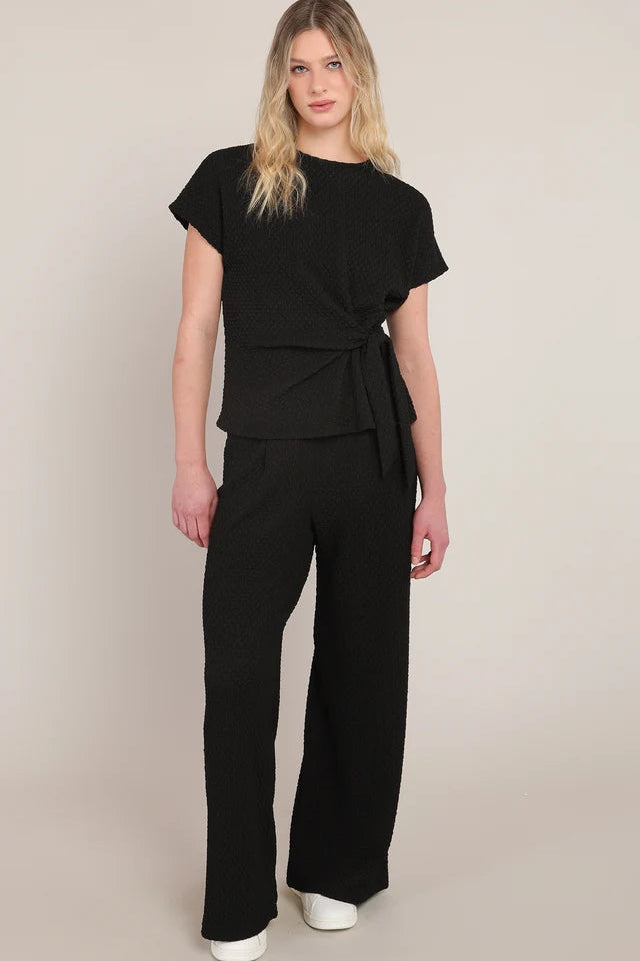A woman modeling a black short-sleeve wrap-style top and matching wide-leg pants against a plain beige studio background.