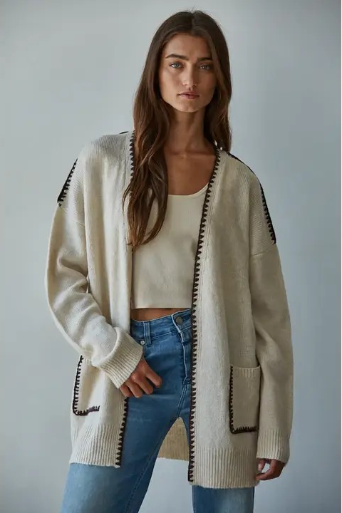 A woman modeling a cream-colored open-front cardigan with black trim details, worn over a white crop top and blue jeans against a plain gray studio background.