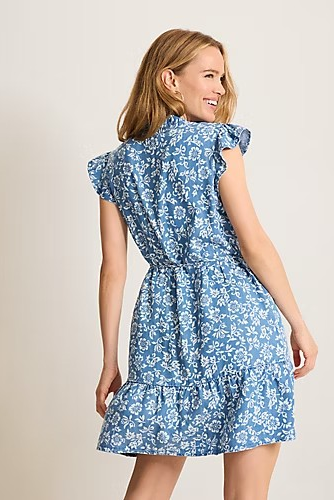 A woman modeling a blue and white floral print dress from the back, featuring ruffled cap sleeves and a ruffled hemline, against a plain beige studio background.