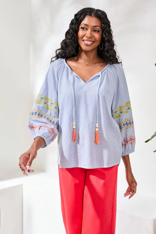 Woman wearing a light blue blouse with colorful patterns and tassels, paired with bright pink pants, against a white background.