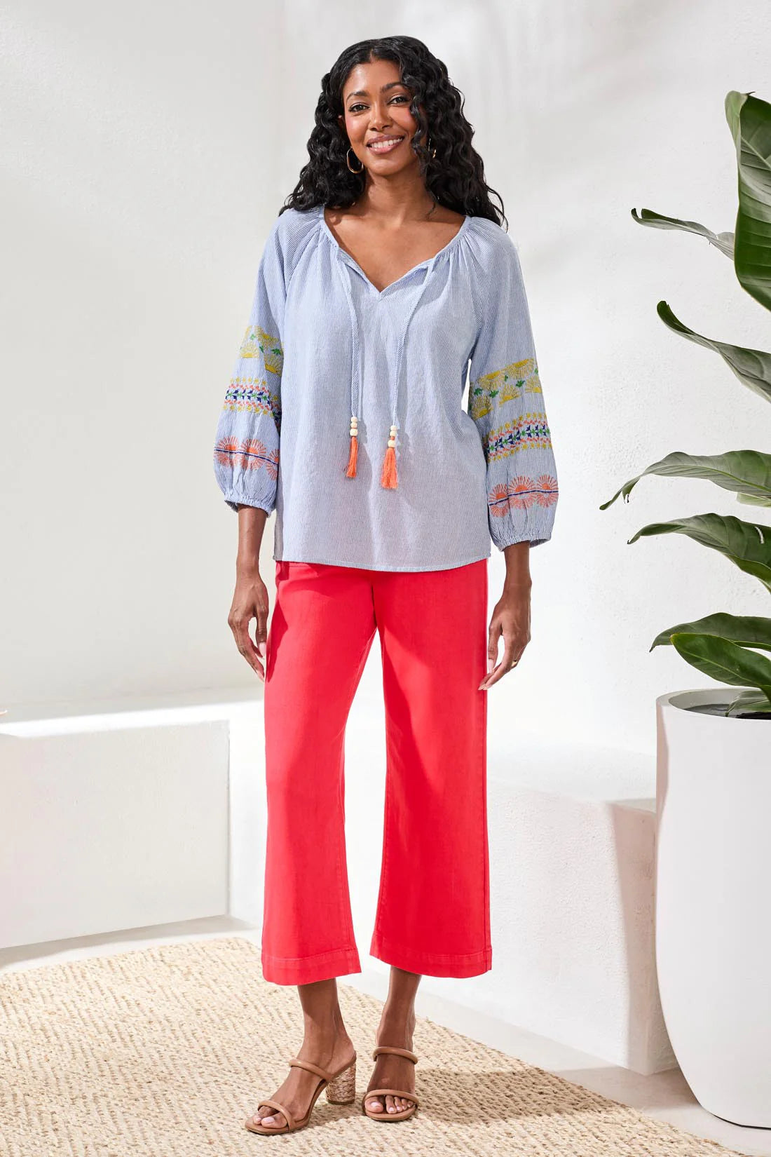 Woman wearing a light blue blouse with colorful sleeves and red pants in a bright room.