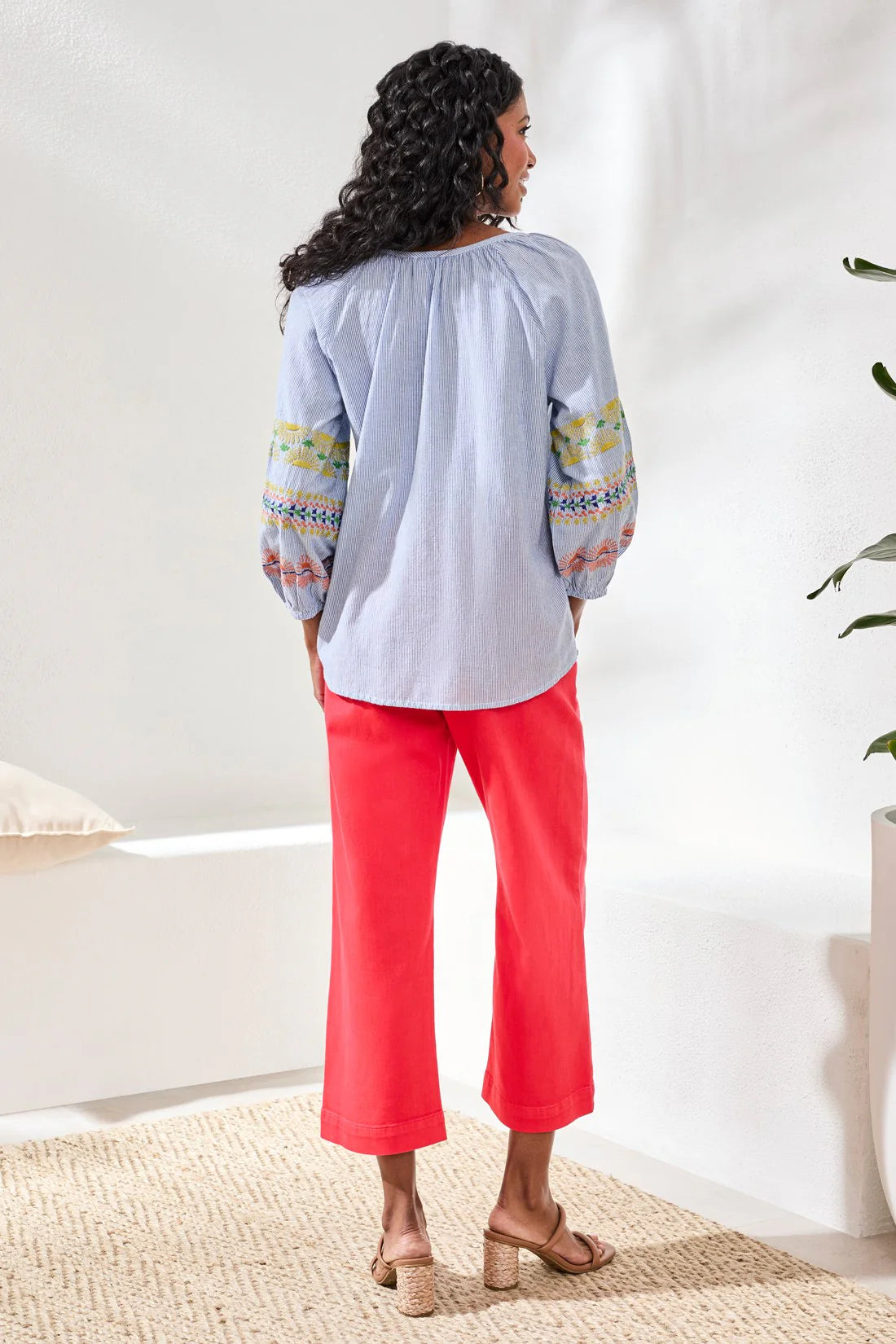 Woman wearing a light blue top with colorful patterns and red pants in a room.