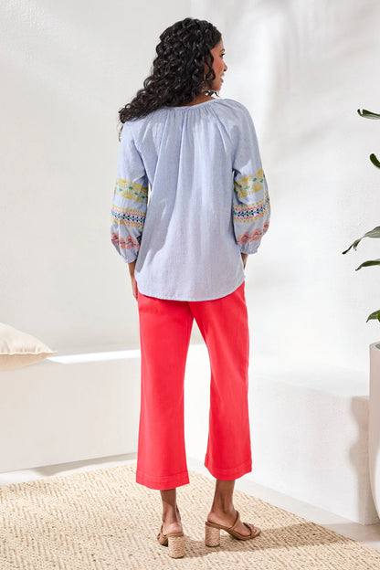 Woman wearing a light blue top with colorful patterns and red pants in a room.