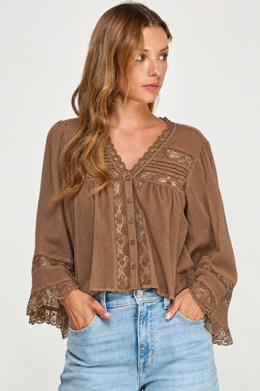 A woman wearing a brown long-sleeve blouse with lace trim and button details, paired with light blue jeans, photographed against a plain light gray background.