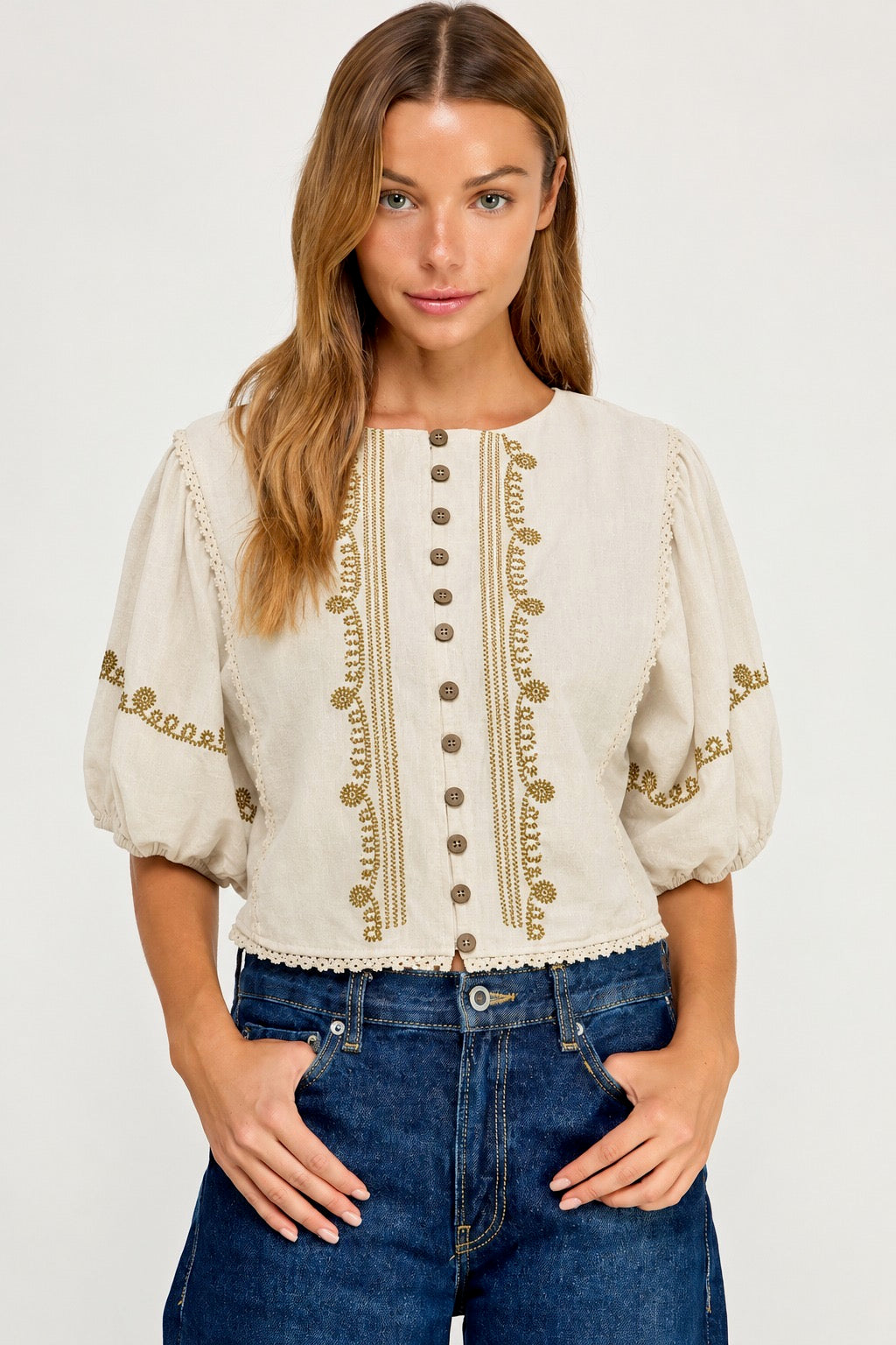 A woman wearing a cream-colored blouse with gold embroidery and button details, paired with blue jeans, against a plain light gray background.