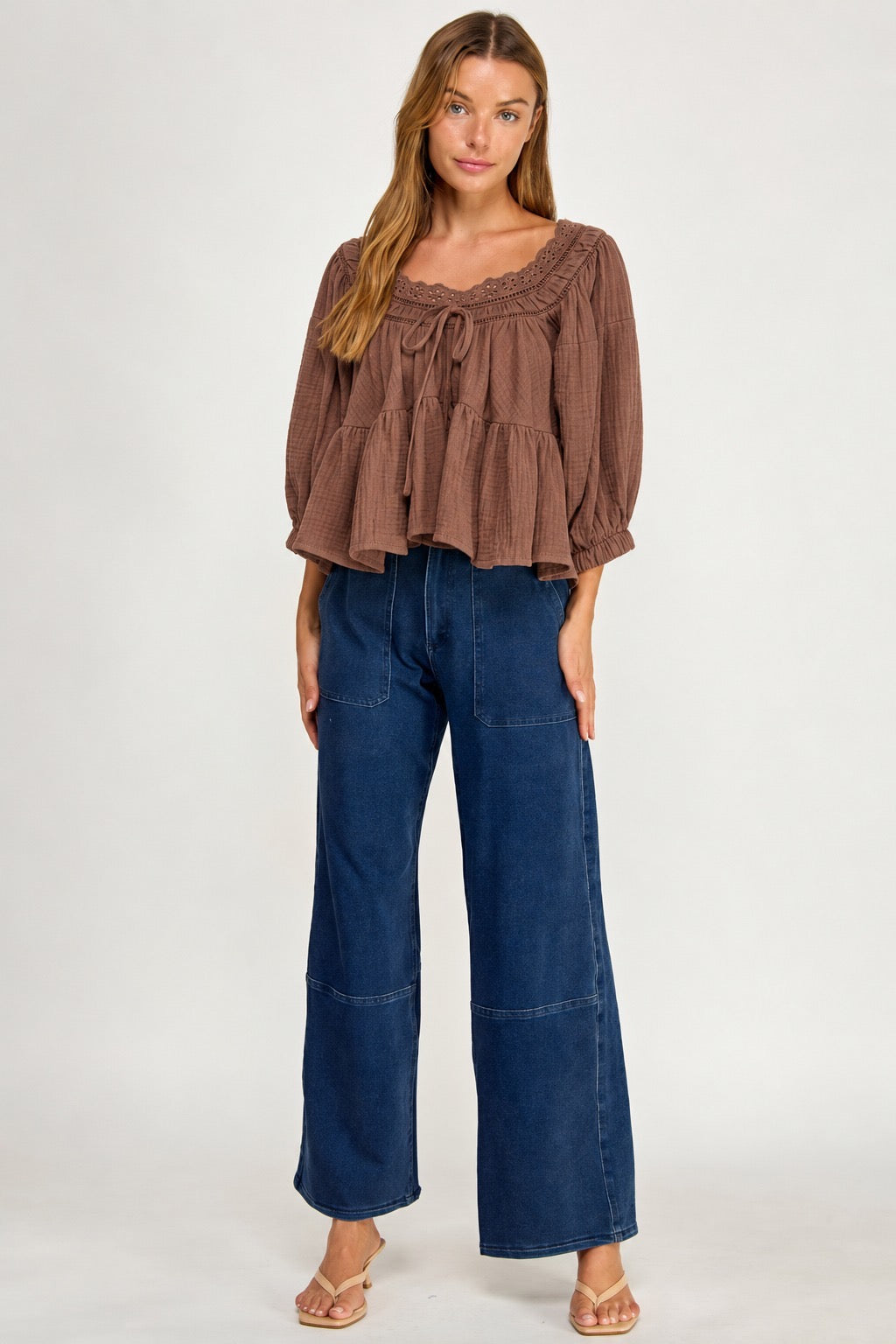 A woman modeling a brown ruffled long-sleeve top paired with dark blue wide-leg jeans and beige sandals, standing against a plain light gray studio background.