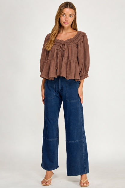 A woman modeling a brown ruffled long-sleeve top paired with dark blue wide-leg jeans and beige sandals, standing against a plain light gray studio background.