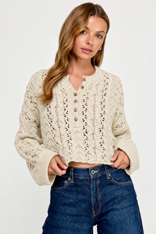 A woman wearing a cream-colored, long-sleeve cable-knit cropped cardigan with a buttoned henley neckline, paired with dark blue jeans, against a plain white studio background.