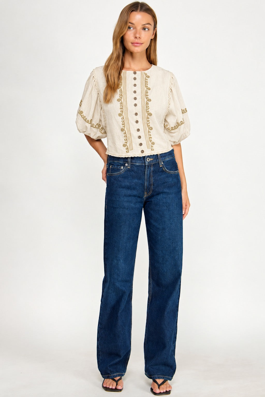 A woman modeling a cream puff-sleeve blouse with gold embroidery and buttons, paired with dark blue wide-leg jeans and black sandals, against a plain white studio background.