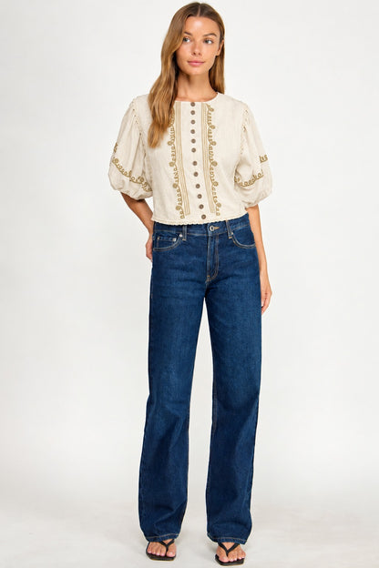 A woman modeling a cream puff-sleeve blouse with gold embroidery and buttons, paired with dark blue wide-leg jeans and black sandals, against a plain white studio background.
