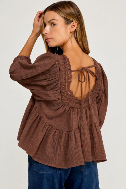 A woman wearing a brown textured long-sleeve blouse with a lace-trimmed open back and tie detail, paired with blue jeans, photographed against a light gray studio background.