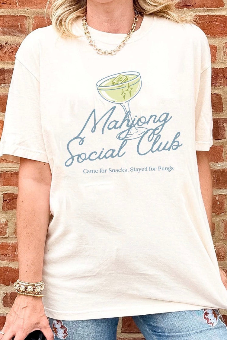 A woman wearing an oversized cream-colored graphic t-shirt with a blue and green 'Mahjong Social Club' design, standing against a red brick wall.