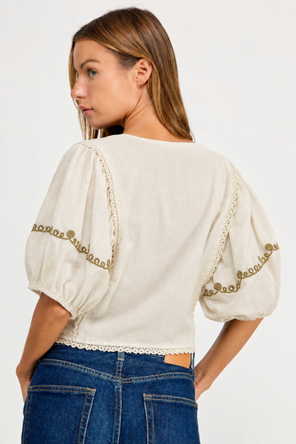 A woman shown from the back wearing a cream-colored puff-sleeve blouse with gold embroidered trim and blue jeans, against a plain white studio background.