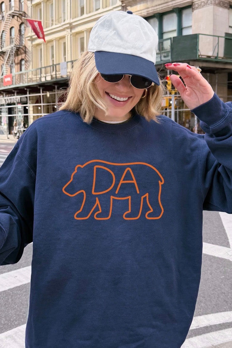 A woman wearing a navy blue sweatshirt with an orange bear outline and the letters 'DA' inside, styled with a white and navy baseball cap, sunglasses, and a coffee cup on a city street.