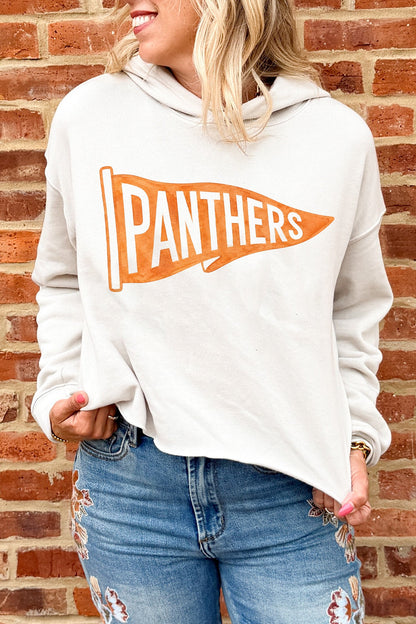 A woman wearing a cream-colored hooded sweatshirt with an orange and white 'PANTHERS' pennant graphic on the front, paired with distressed blue jeans, standing against a red brick wall.