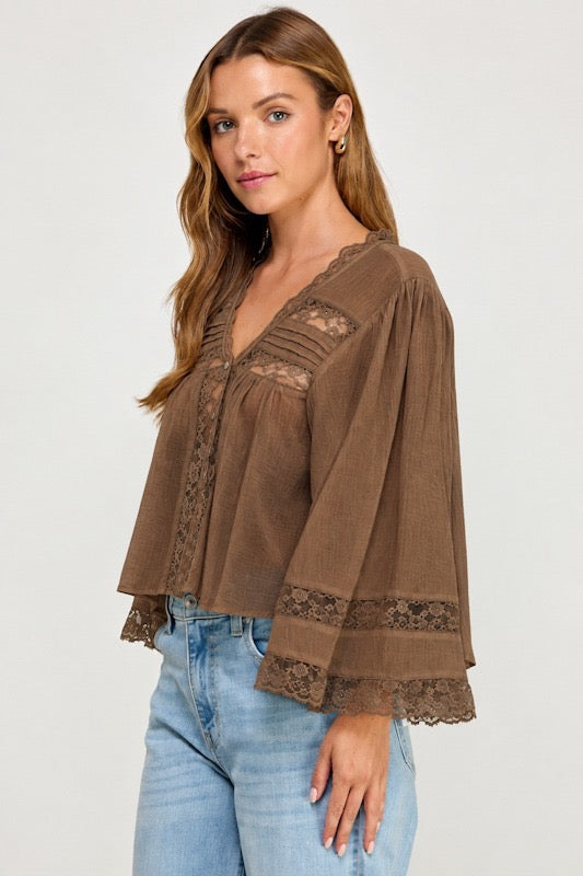 A woman wearing a brown lace-trimmed blouse with bell sleeves and light blue jeans, photographed against a plain light gray studio background.