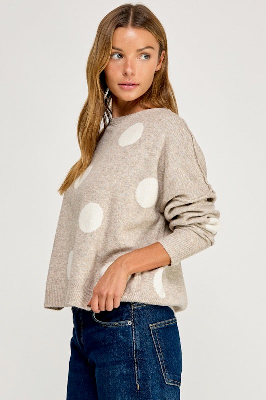A woman wearing a light beige knit sweater with large white polka dots and dark blue jeans, photographed against a plain light gray studio background.