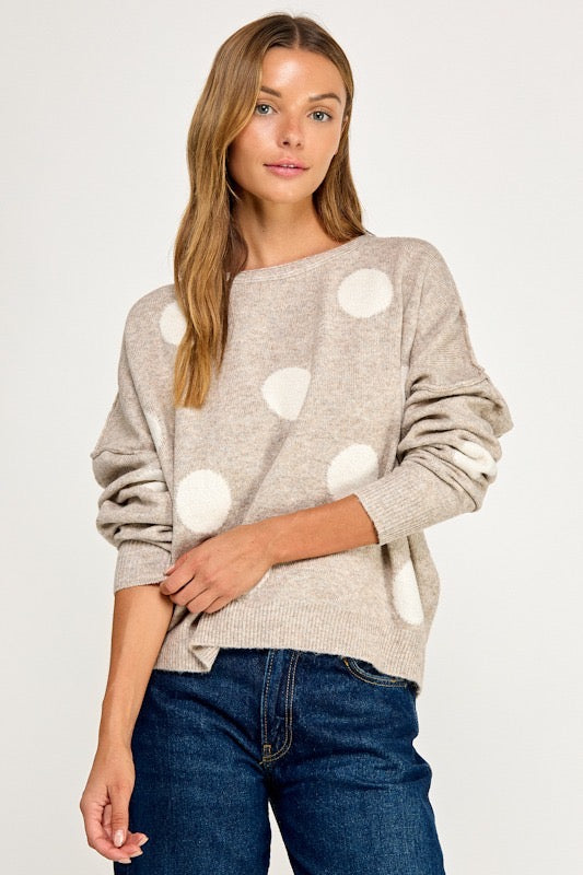 A woman wearing a light beige knit sweater with large white polka dots and dark blue jeans, photographed against a plain light gray studio background.