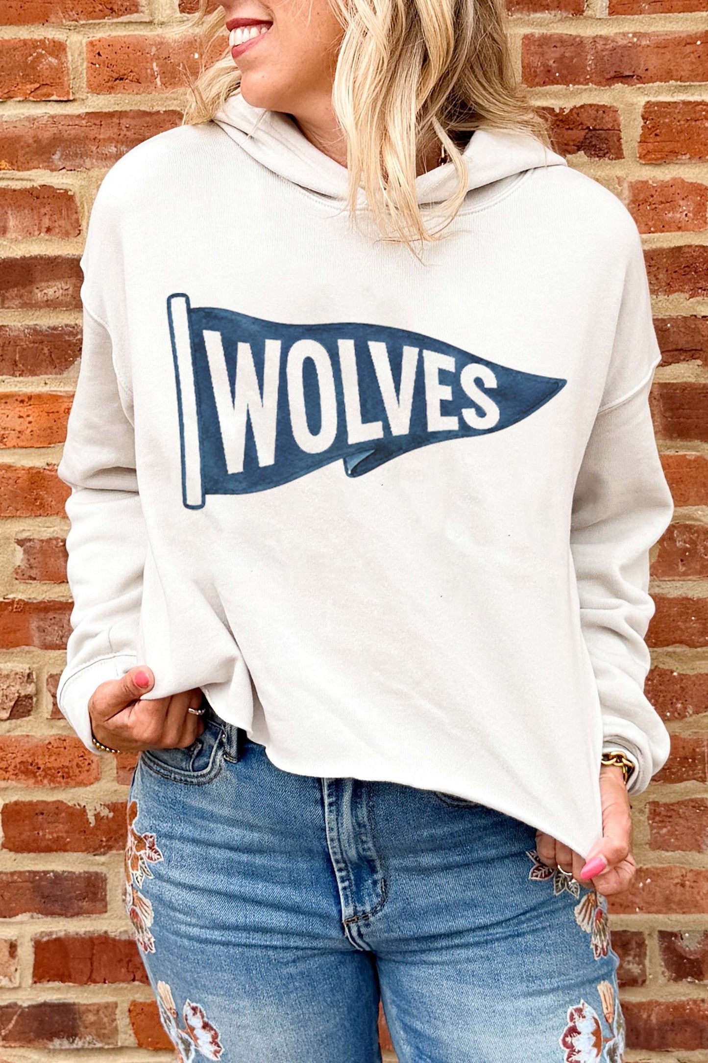 A woman wearing a light beige hoodie with a navy blue and white 'WOLVES' pennant graphic on the front, paired with distressed blue jeans, standing against a red brick wall.