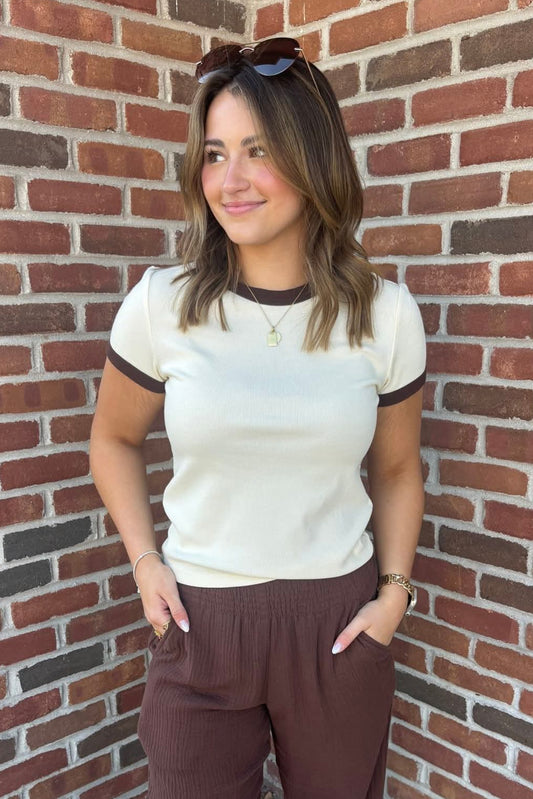 Woman wearing a white shirt with black trim and brown pants standing against a brick wall.