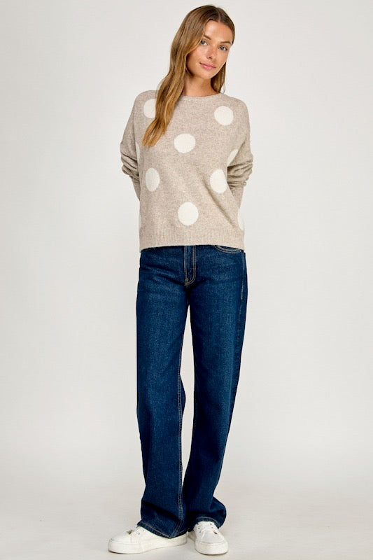 A woman modeling a beige crewneck sweater with large white polka dots, paired with dark blue jeans and white sneakers, against a plain light gray studio background.