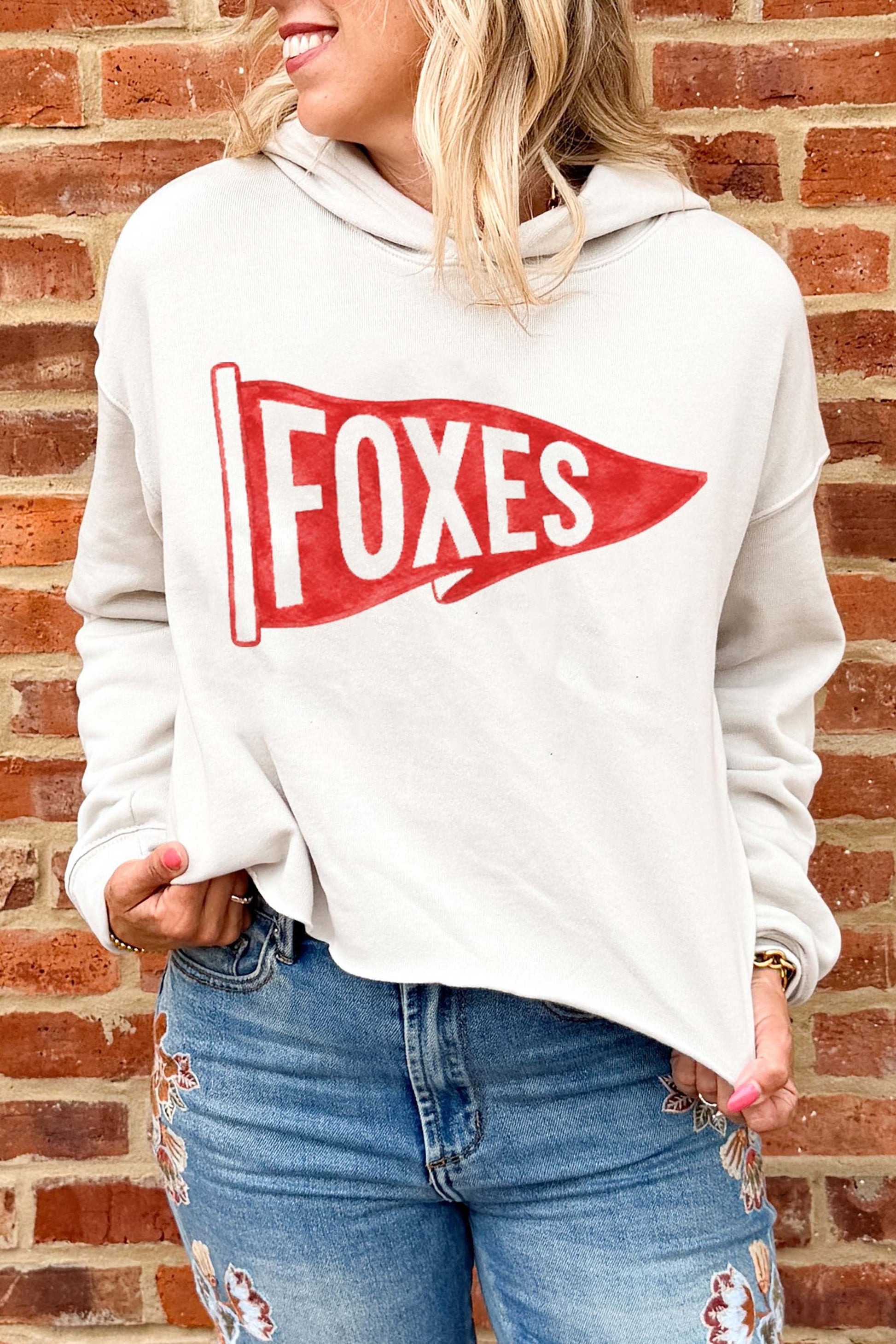 A woman wearing a light beige hoodie with a red and white 'FOXES' pennant graphic, paired with distressed blue jeans, standing against a red brick wall.