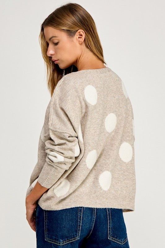 A woman wearing a light beige knit sweater with large white polka dots, shown from a side-back angle against a plain light gray studio background.