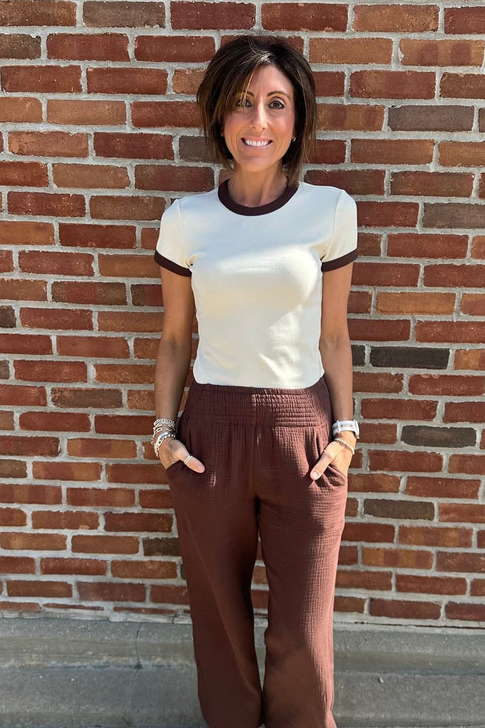 Woman standing in front of a brick wall wearing a white and black top and brown pants.