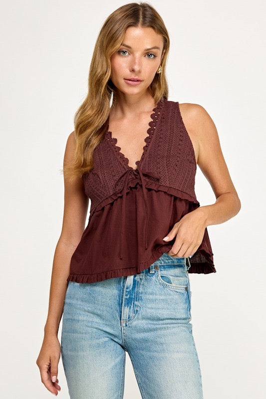 A woman modeling a sleeveless dark burgundy top with a scalloped lace V-neckline, ruffled tie detail, and tiered peplum hem, paired with light-wash blue jeans against a plain white studio background.