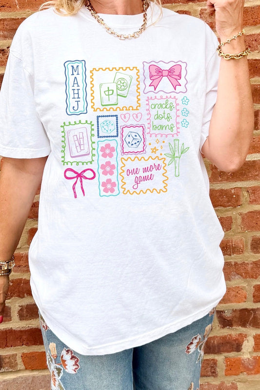 A woman wearing a white graphic t-shirt with a colorful patchwork design of stamps, bows, flowers, and text, paired with distressed blue jeans, standing against a red brick wall.