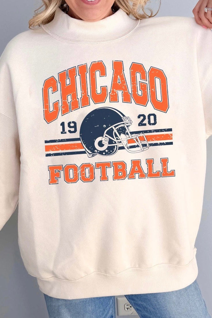 A woman wearing a cream-colored sweatshirt with a distressed Chicago Football graphic on the front, paired with blue jeans, against a solid gray background.
