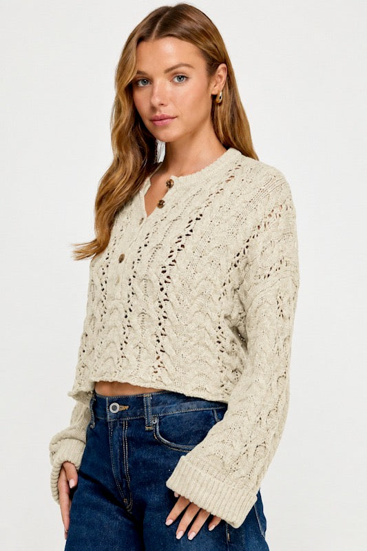A woman modeling a cropped, long-sleeve cable-knit cardigan in cream with brown buttons, paired with dark blue jeans, against a plain white studio background.