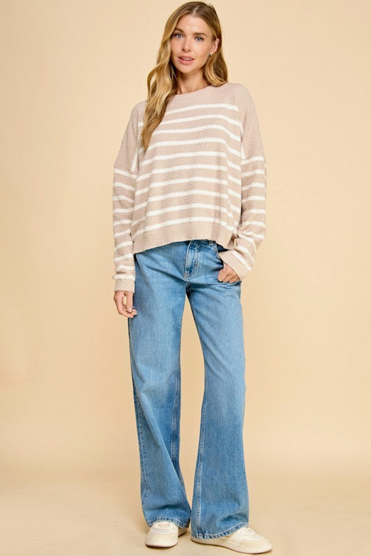 A woman modeling a beige and white horizontally striped long-sleeve knit sweater with blue wide-leg jeans and white sneakers, standing against a solid beige studio background.