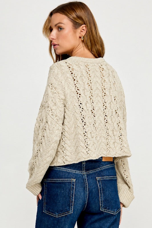 A woman shown from the back wearing a cream cable-knit cropped sweater and blue jeans against a plain light gray studio background.