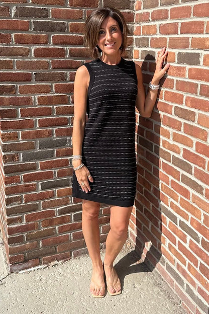 Woman in a black dress standing against a brick wall on a sunny day.