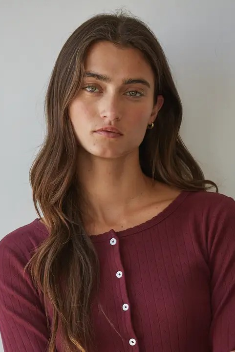 A woman with long brown hair wearing a ribbed long-sleeve henley top in burgundy, standing against a plain light gray wall.