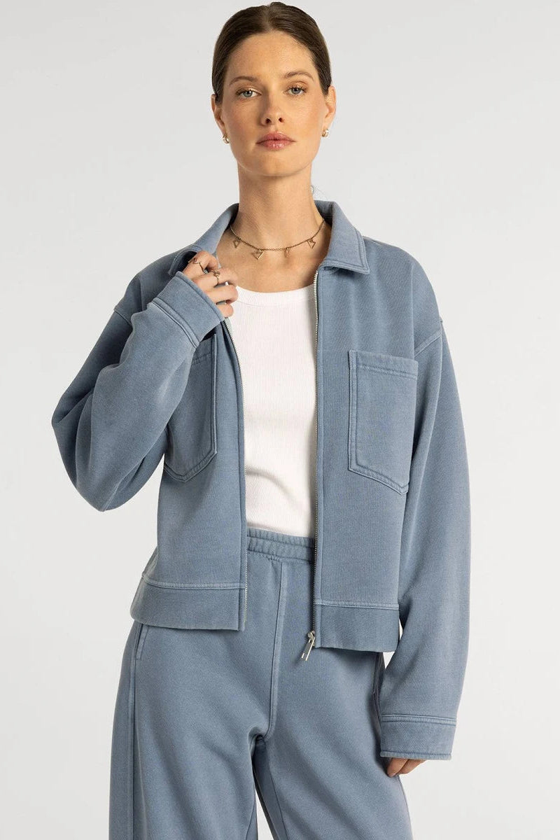 A woman modeling a light blue cropped jacket and matching sweatpants over a white top, photographed against a plain light gray studio background.