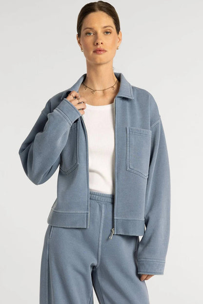 A woman modeling a light blue cropped jacket and matching sweatpants over a white top, photographed against a plain light gray studio background.