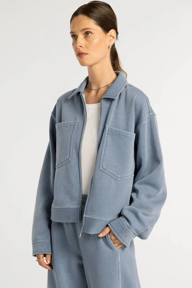 A woman wearing a light blue cropped jacket with front pockets over a white top and matching blue pants, photographed against a plain light gray studio background.