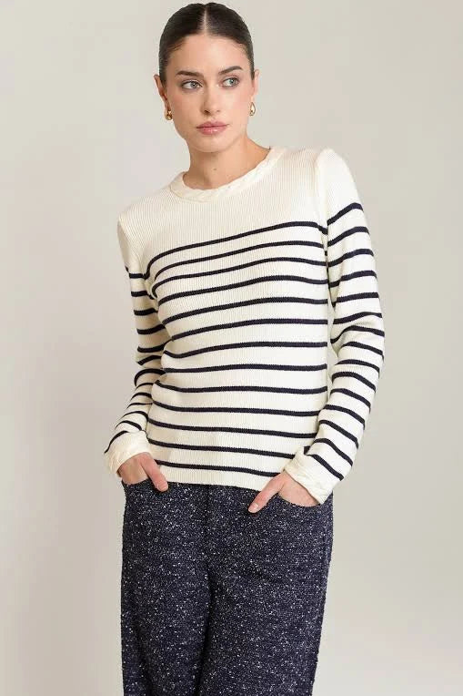 A woman modeling a cream knit sweater with black horizontal stripes across the chest and sleeves, paired with dark speckled pants, against a plain light gray studio background.