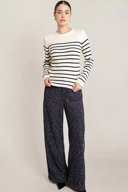 A woman modeling a long-sleeve knit sweater with black and white horizontal stripes, paired with dark wide-leg pants, against a plain light beige studio background.