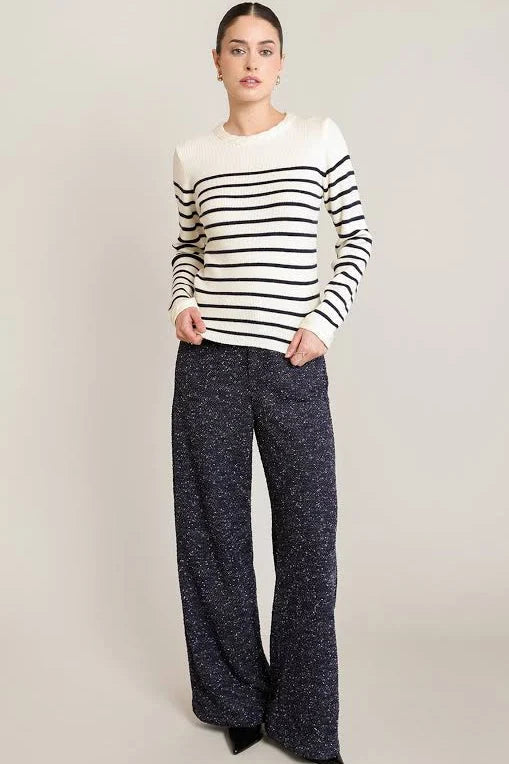A woman modeling a long-sleeve knit sweater with black and white horizontal stripes, paired with dark wide-leg pants, against a plain light beige studio background.