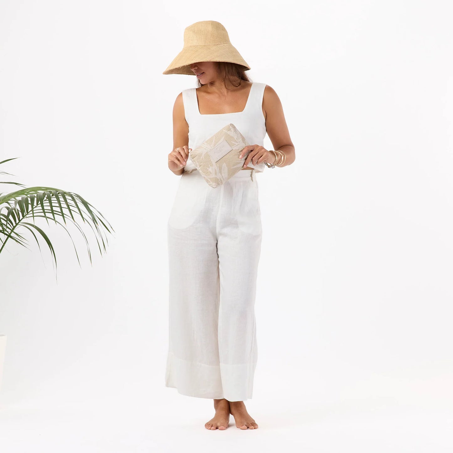 Woman wearing a white sleeveless top and wide-leg pants, holding a beige bag, on a white background.