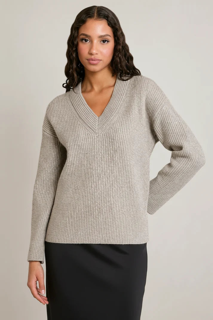 A woman modeling a light beige ribbed knit sweater with a V-neck and long sleeves, paired with a black skirt, against a plain light gray studio background.