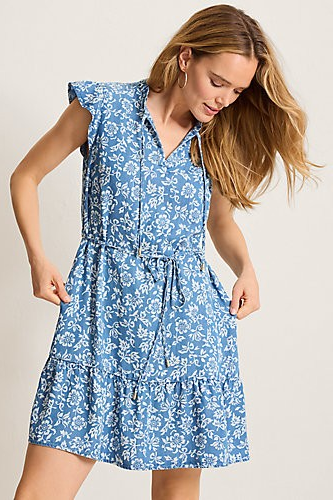 A woman wearing a sleeveless blue and white floral print dress with a tiered skirt, shown against a plain light beige studio background.